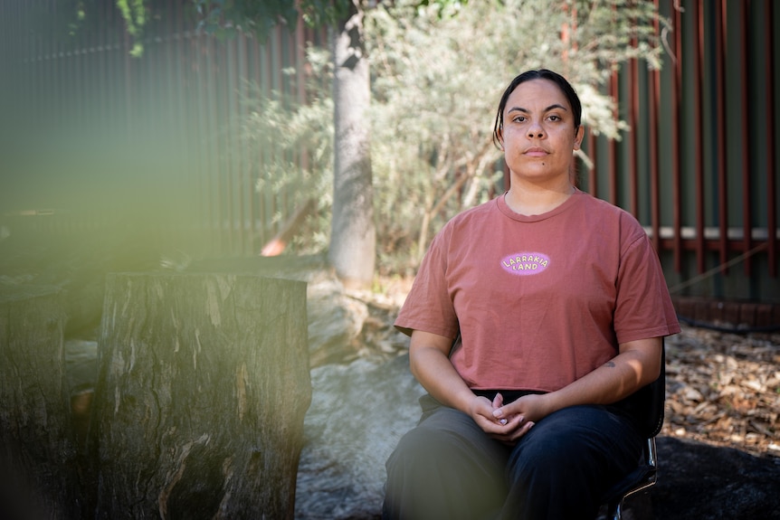 a young aboriginal woman wearing a pink t-shirt sitting on a chair in front of shrubs