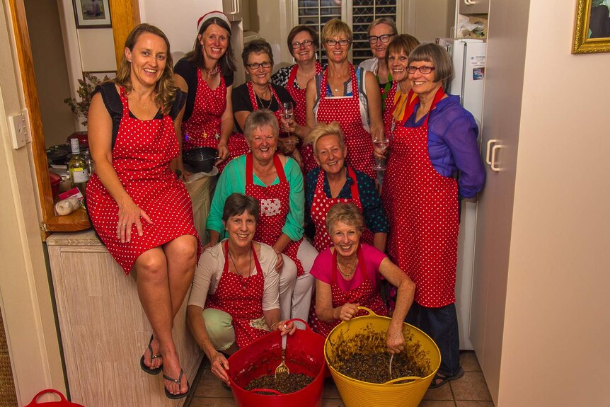 Women in red spotted aprons sit or stand in a kitchen in front of two laundry tubs of pudding mix