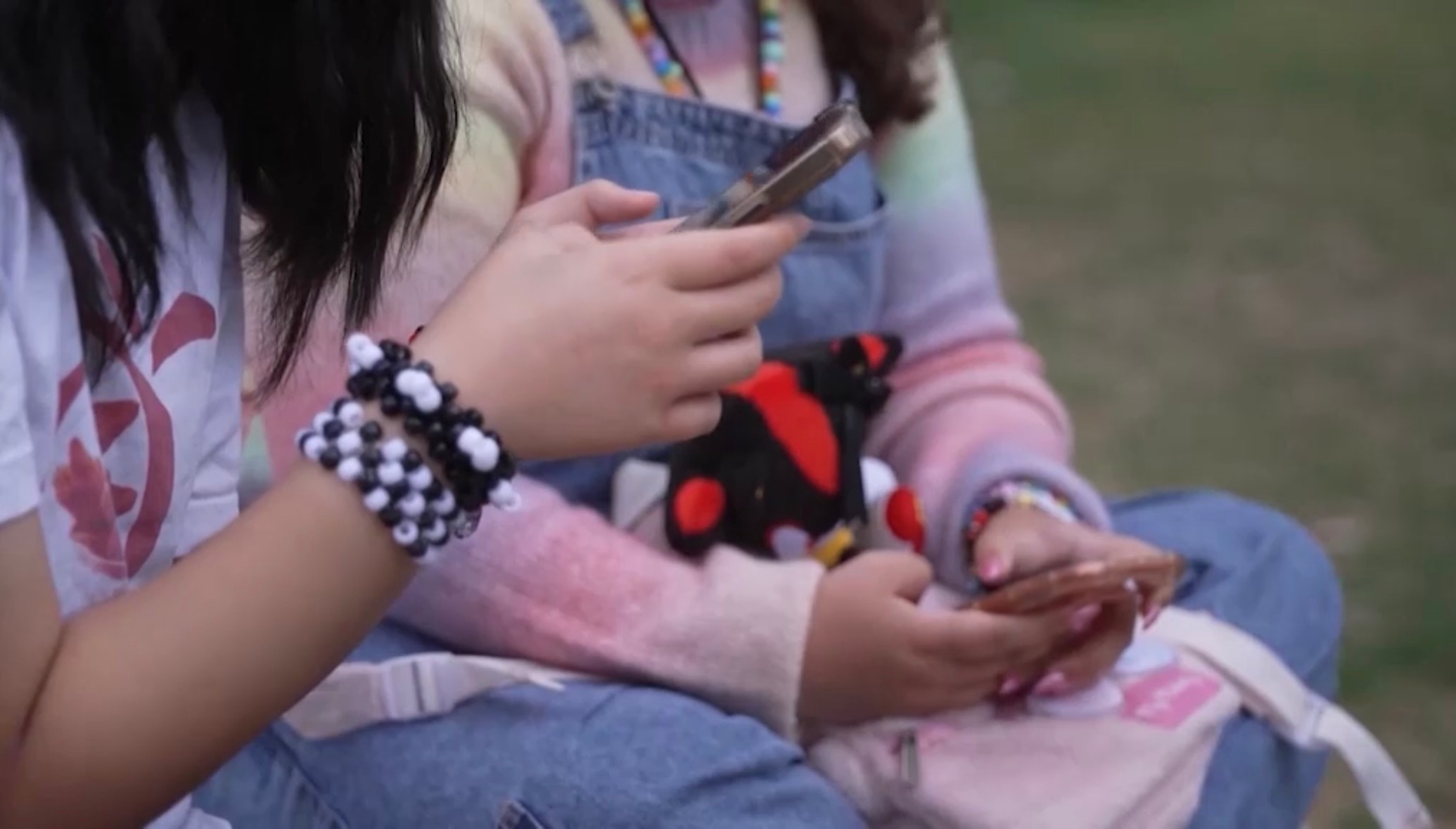 Two girls look at mobile phones. One wears black and white beads on her wrist. The other wears overalls and a colourful jumper.