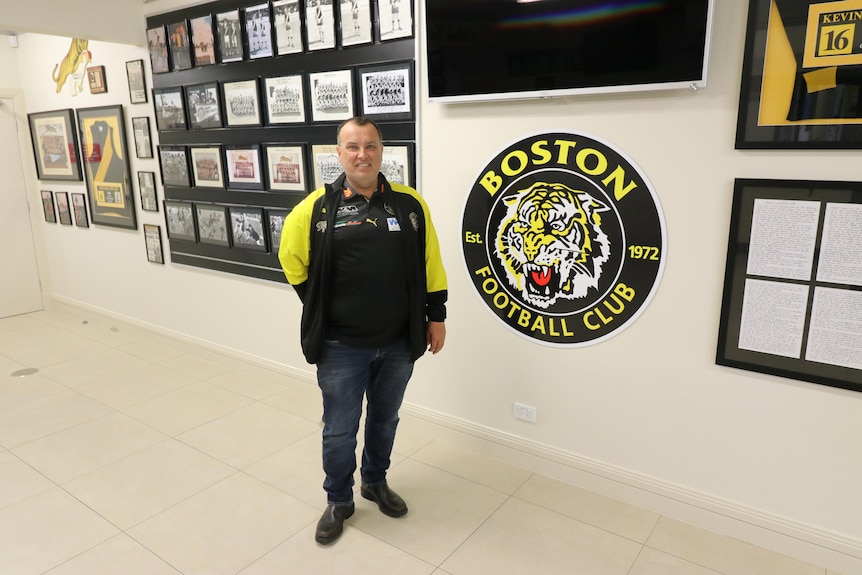 A man, wearing a black and yellow jumper, stands in front of honour boards at a football club.