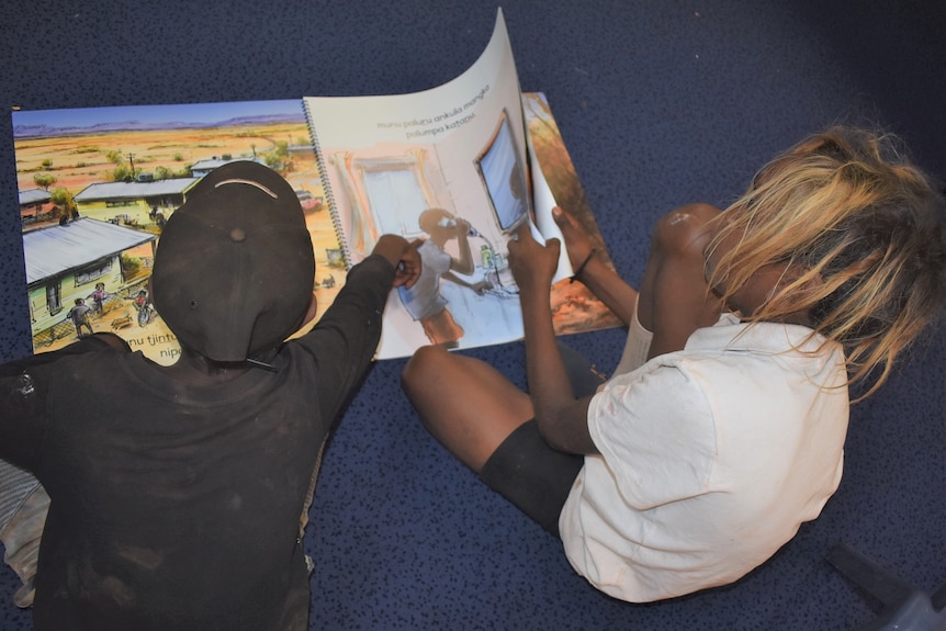 Two young boys sit cross-legged on a blue carpet flipping through and pointing at a children's book