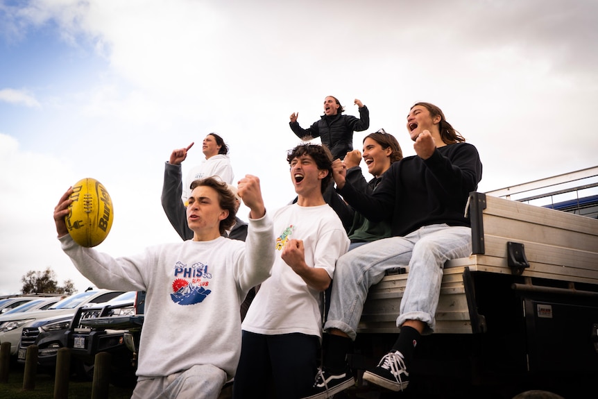 A group of teenage boys cheer on football teams from the back of a ute.