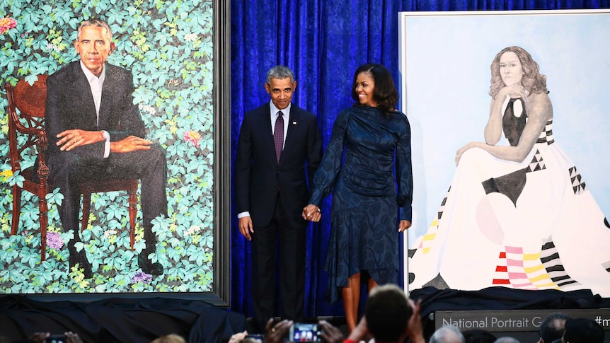 Barack Obama and Michelle Obama hold hands and stand in between their portraits.