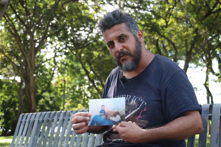 A man sitting on a bench in front of trees, looking serious and holding a photo of himself and a small boy.