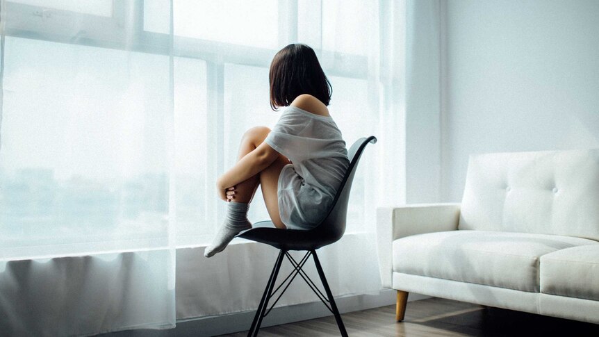 A young woman with dark hair sits on a chair while staring out the window.