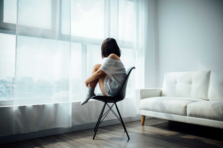 A young woman with dark hair sits on a chair while staring out the window.