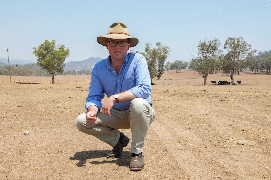 A man wearing a hat squats down in a drought-affected, dry paddock