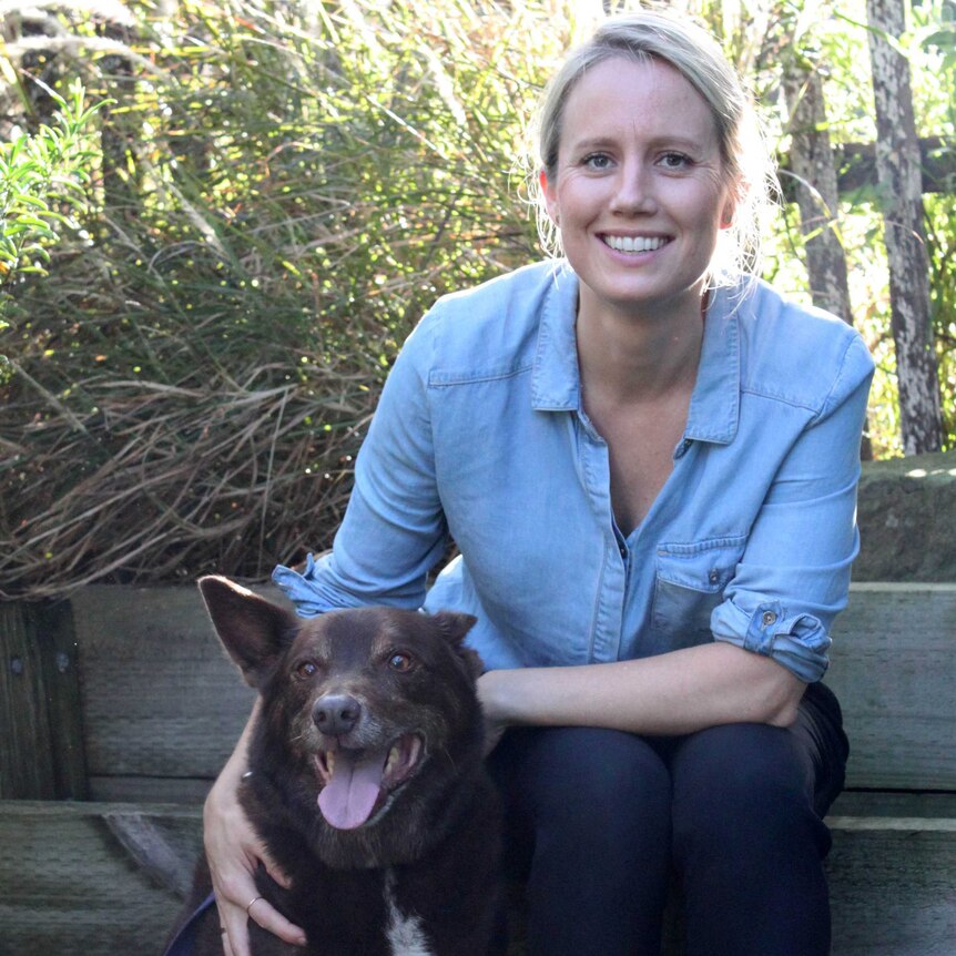 A smiling woman in a blue shirt with her hand on a brown kelpie dog.