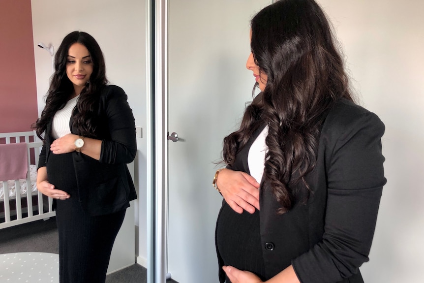 A young white woman with long dark hair looking into a full-body mirror, holding her stomach