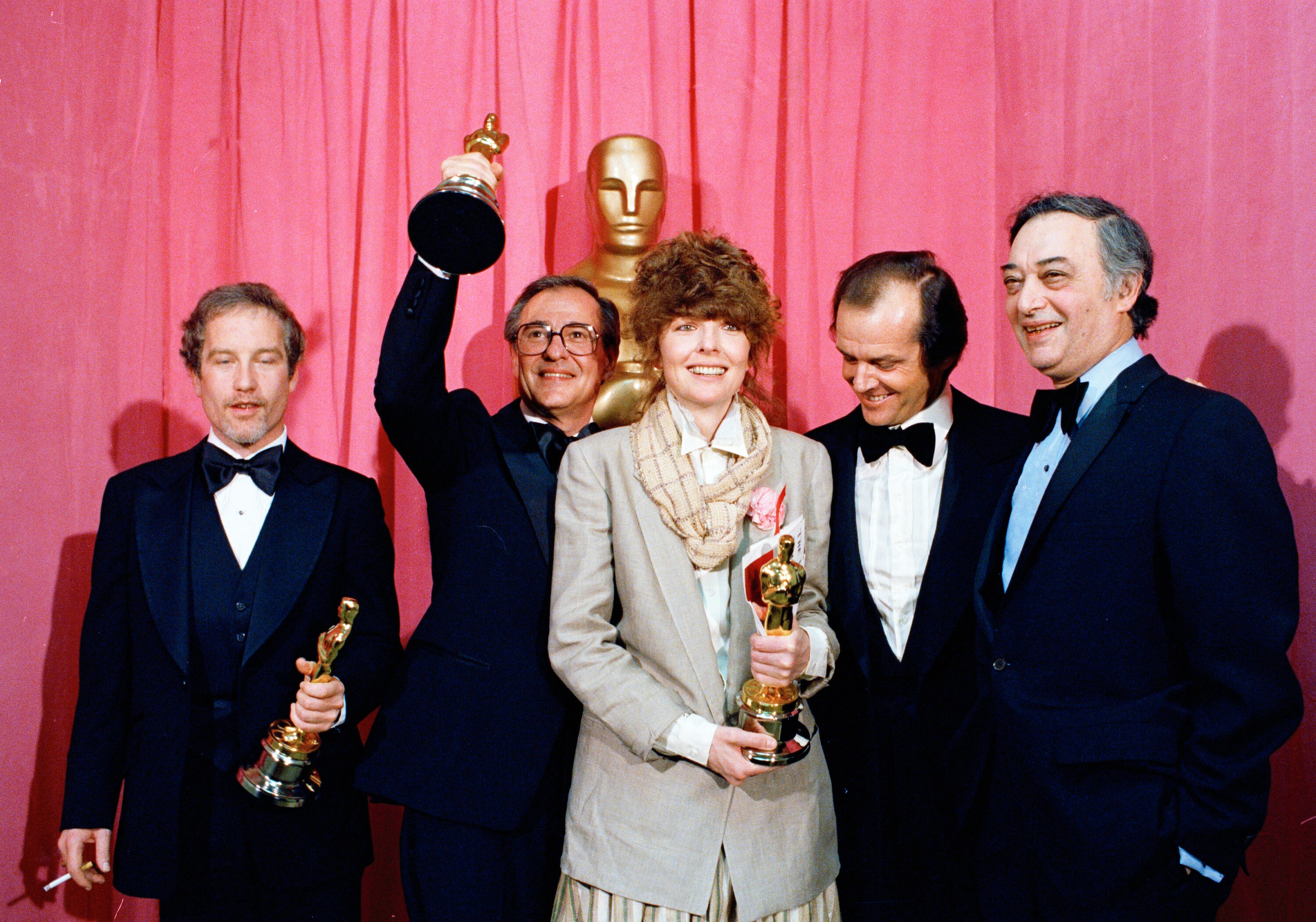 Diane Keaton (centre) in a grey suit jacket and checked scarf holding an oscar, surrounded by men in suits. 