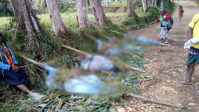 On the side of a dirt road, bodies are pictured blurred in blue mourning wraps tied to logs, as three people watch over them.