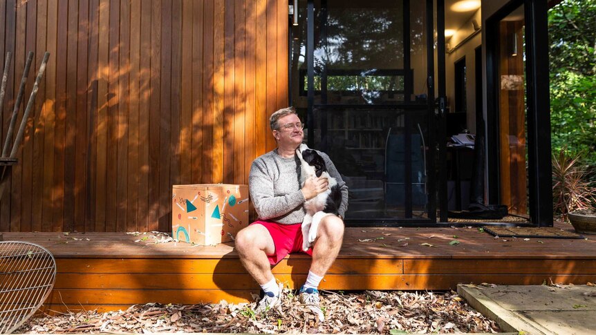 A man in a grey jumper and red shorts sits on the front porch of a modern wood home hugging a border collie.