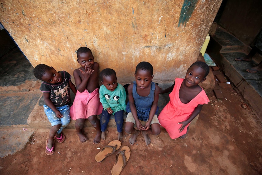 A shit looking down on five small children dressed in bright clothes and smiling at the camera sitting on stone on a dirt road.