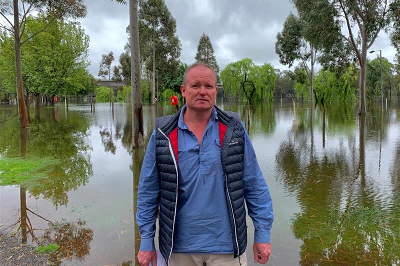 Man in long-sleeve shirt and vest standing in front of flooded caravan park at Murray River