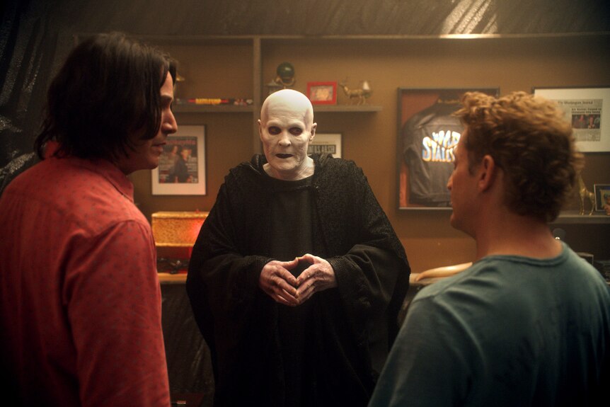 In a room with frame music memorabilia, a robe wearing person with white ghoulish appearance stands chatting to two men.