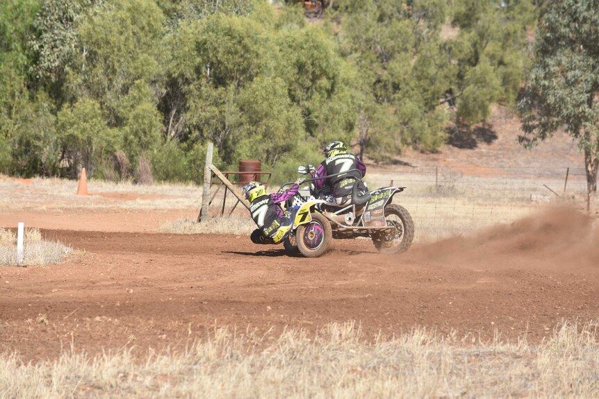 Two men on motorbike and sidecar race around a dirt track.  The one in the sidecar is leaning out of it.