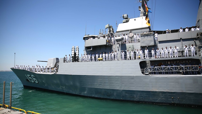 Dozens of servicemen and women line the sides of a large white naval vessel docked in a harbour