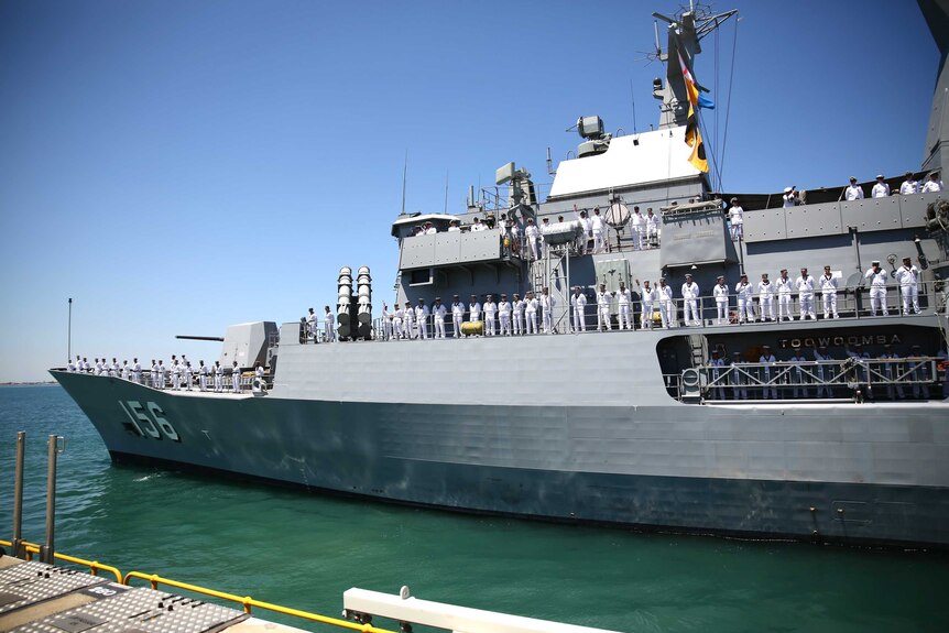 Dozens of servicemen and women line the sides of a large white naval vessel docked in a harbour