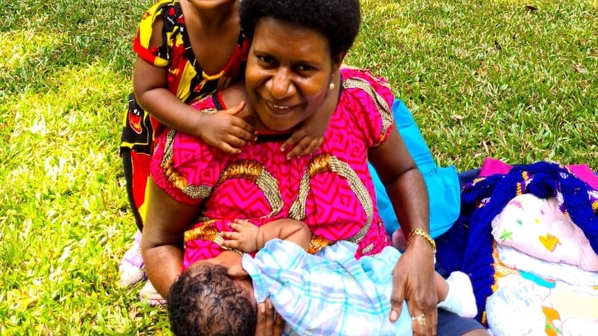 A woman sits on the grass in PNG breastfeeding her baby with a small girl behind her