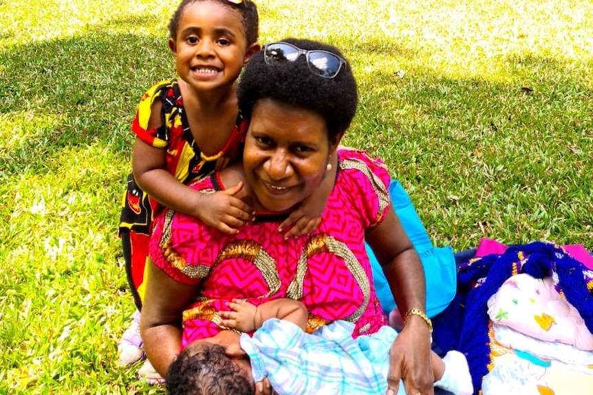 A woman sits on the grass in PNG breastfeeding her baby with a small girl behind her