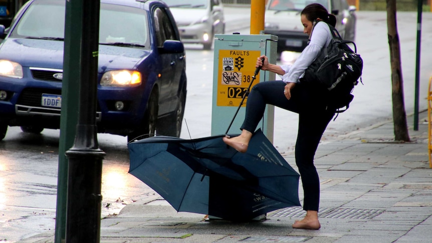 A woman in a white shirt hold a large broken umbrella upside down and tries to straighten it with her foot.