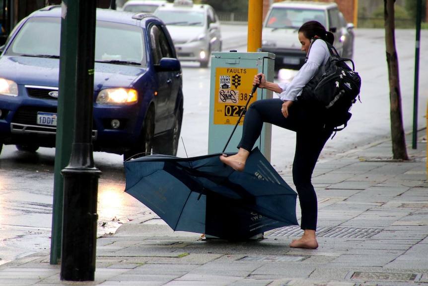 A woman in a white shirt hold a large broken umbrella upside down and tries to straighten it with her foot.