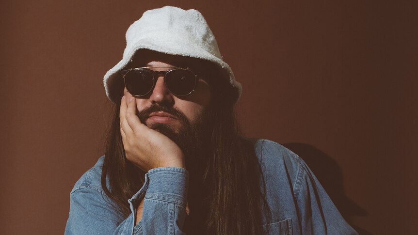 Matthew E. White wear a towel hat and sunglasses. He has his head in one hand. He wears a denim shirt.