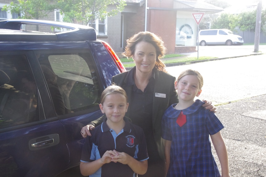 Two kids in thir school uniform in front of a car with a lady.