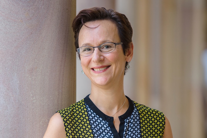 A woman with short hair, wearing glasses, leaning on a column of a building.