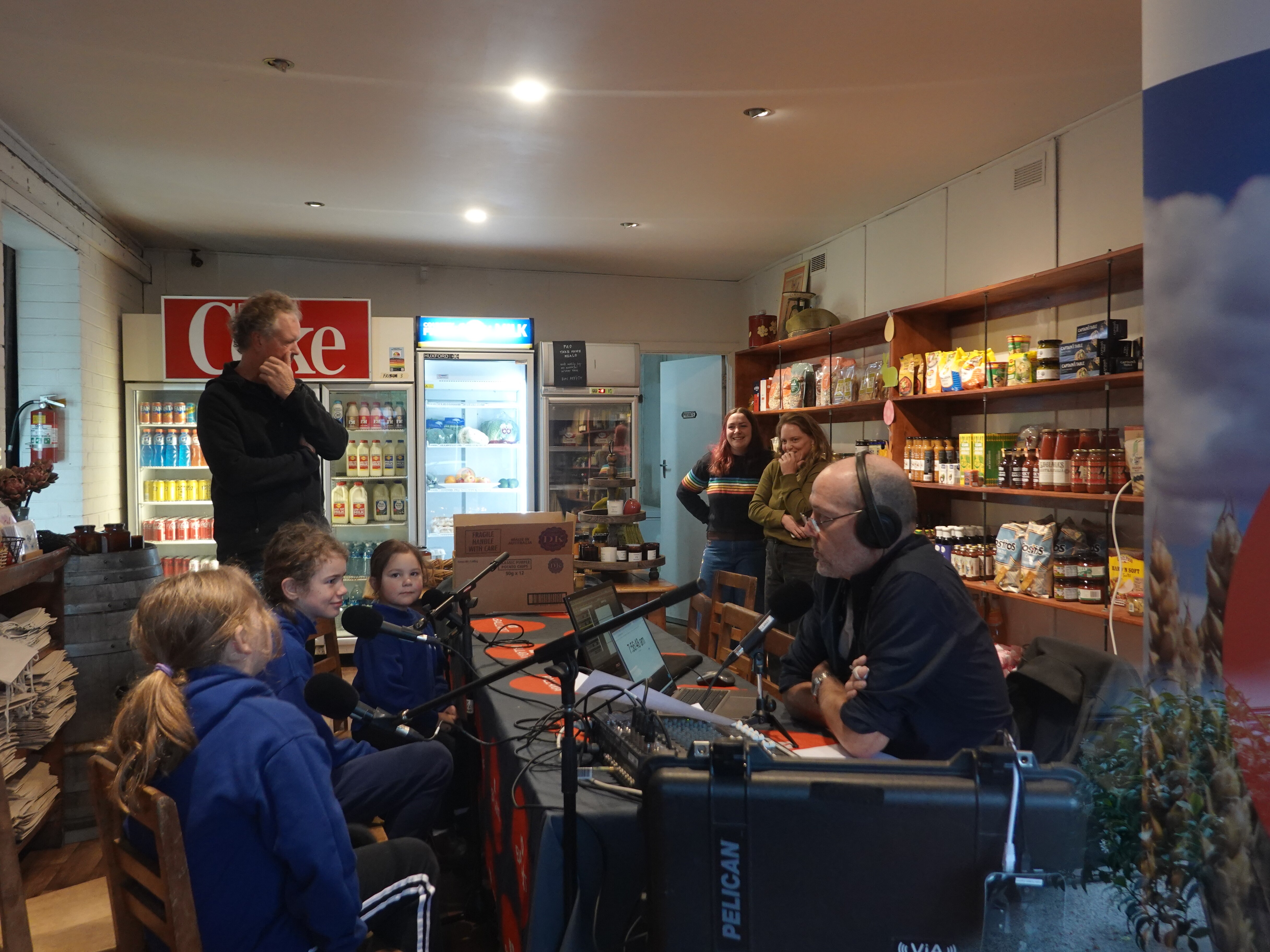 Kids and an ABC presenter sit at a table in a general store and talk into microphones.