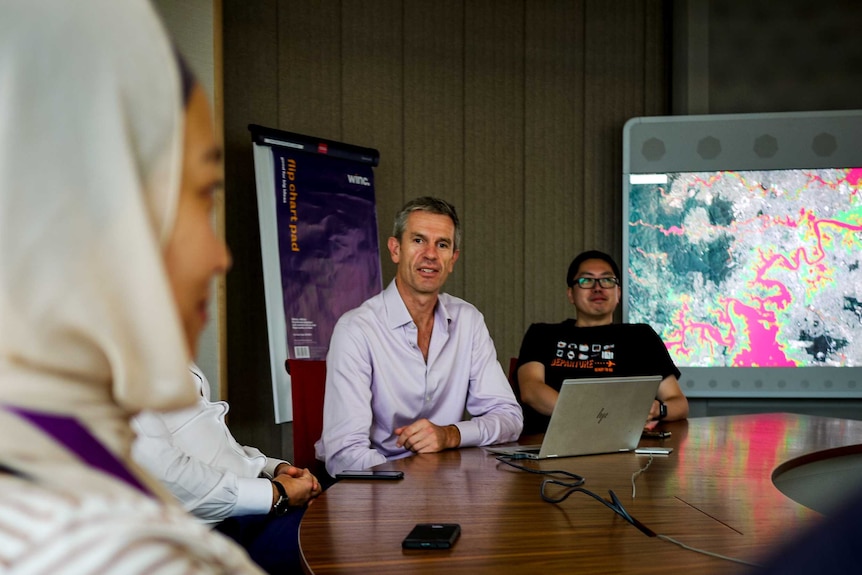 A group of people sitting at circular meeting table, woman in foreground, one man looking at camera, one man on laptop