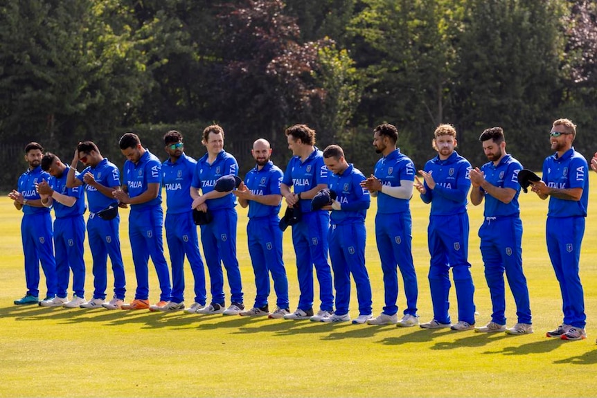 13 men stand on grass wearing blue pants and shirts