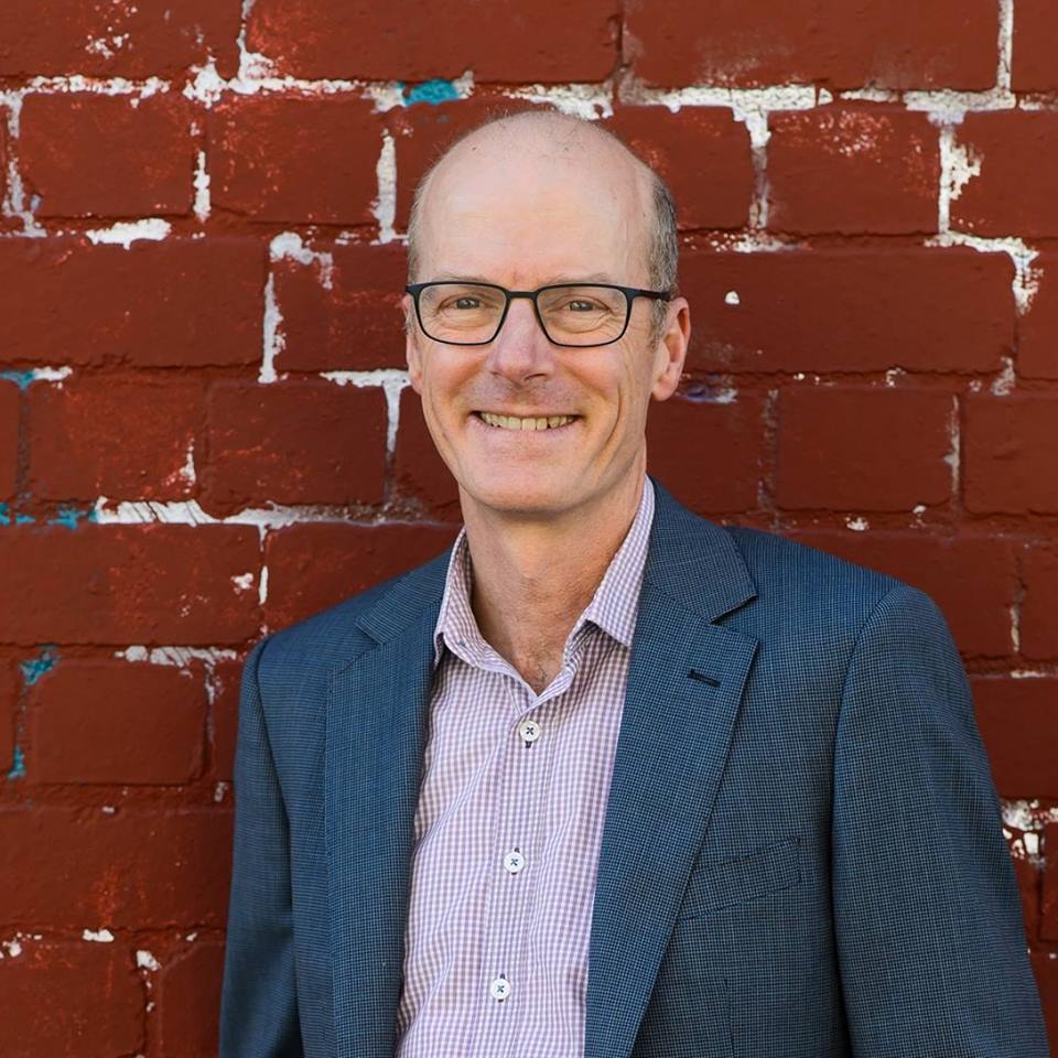 A man in spectacles stands in front of a red brick wall.