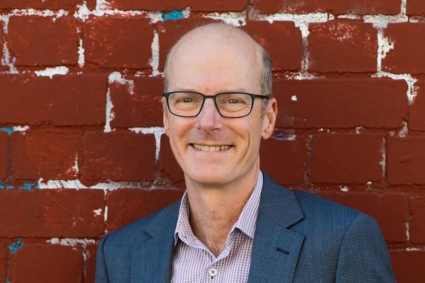 A man in spectacles stands in front of a red brick wall.