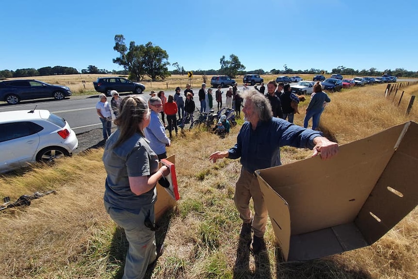 Around 20 people gather on a roadside, in front of a vacant paddock to protest a proposed development