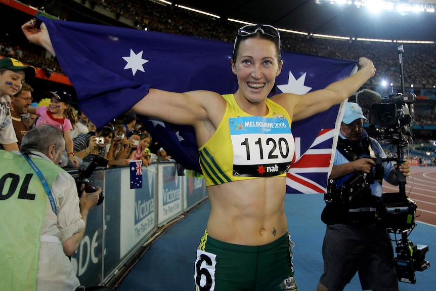 Jana Pittman, in green and gold lycra top and bottom, smiles widely holding the outstretched Australian flag, with crowd behind.