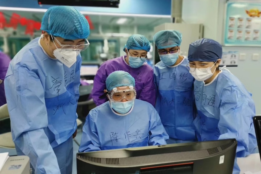 Medical workers in Wuhan wearing masks and blue goggles who have written their names on their scrubs, stare at a computer.