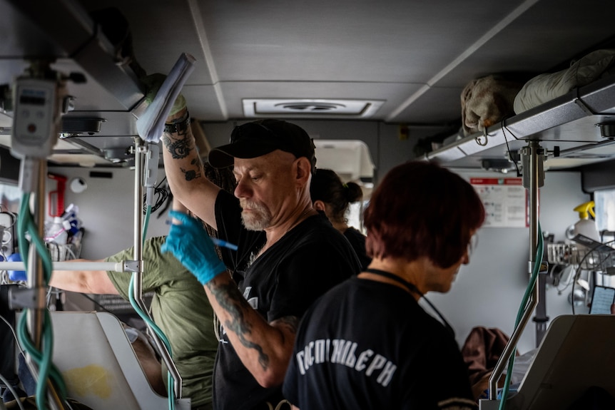 Doctors inside a bus equipped with hospital beds. One man wearing a blue surgical glove over his tattooed hands is in focus
