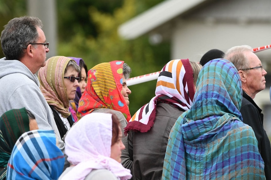A group of people, some covering their heads, stand together outside a mosque to observe a ceremony