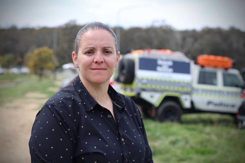 A woman stands facing the camera in front of bushland and a police search and rescue vehicle.