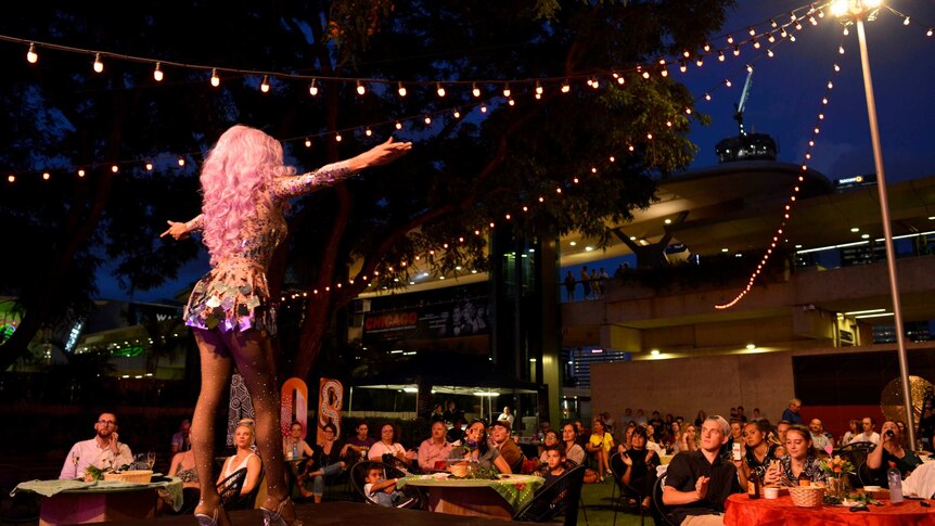 The back view of a drag queen in a pink wig and glittery jumpsuit on stage and looking out on a crowd.