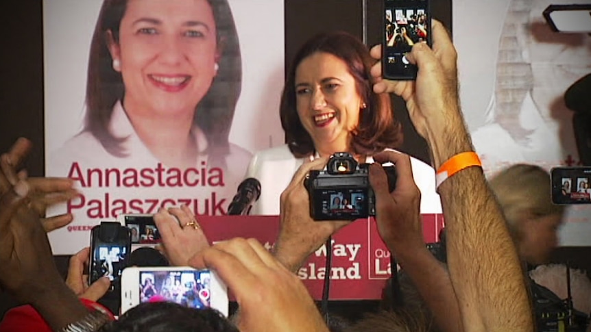 A woman stands at a lectern smiling in front of posters of herself