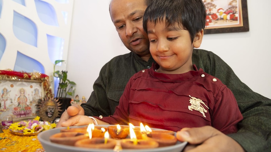 A balding man in a green kurta and a smiling 4yo boy in red t-shirt hold a plat of clay lamps.