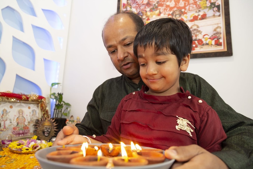 A balding man in a green kurta and a smiling 4yo boy in red t-shirt hold a plat of clay lamps.