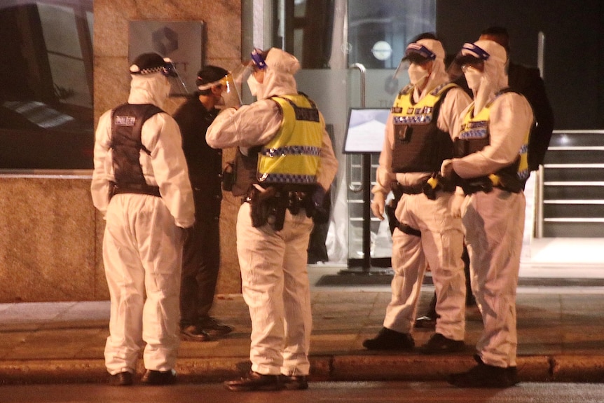 A group of four police officers in white overalls, wearing face masks and shields, outside a building.