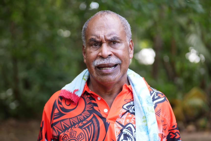 Torres Strait Islander man with orange shirt and gray mustache looks into camera. He has a towel around his neck.