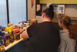 A woman holds a baby and daughter, with her back to the camera facing a table full of groceries