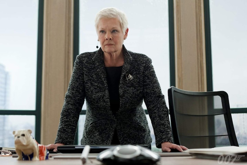 A woman leans on an office desk with a serious expression.