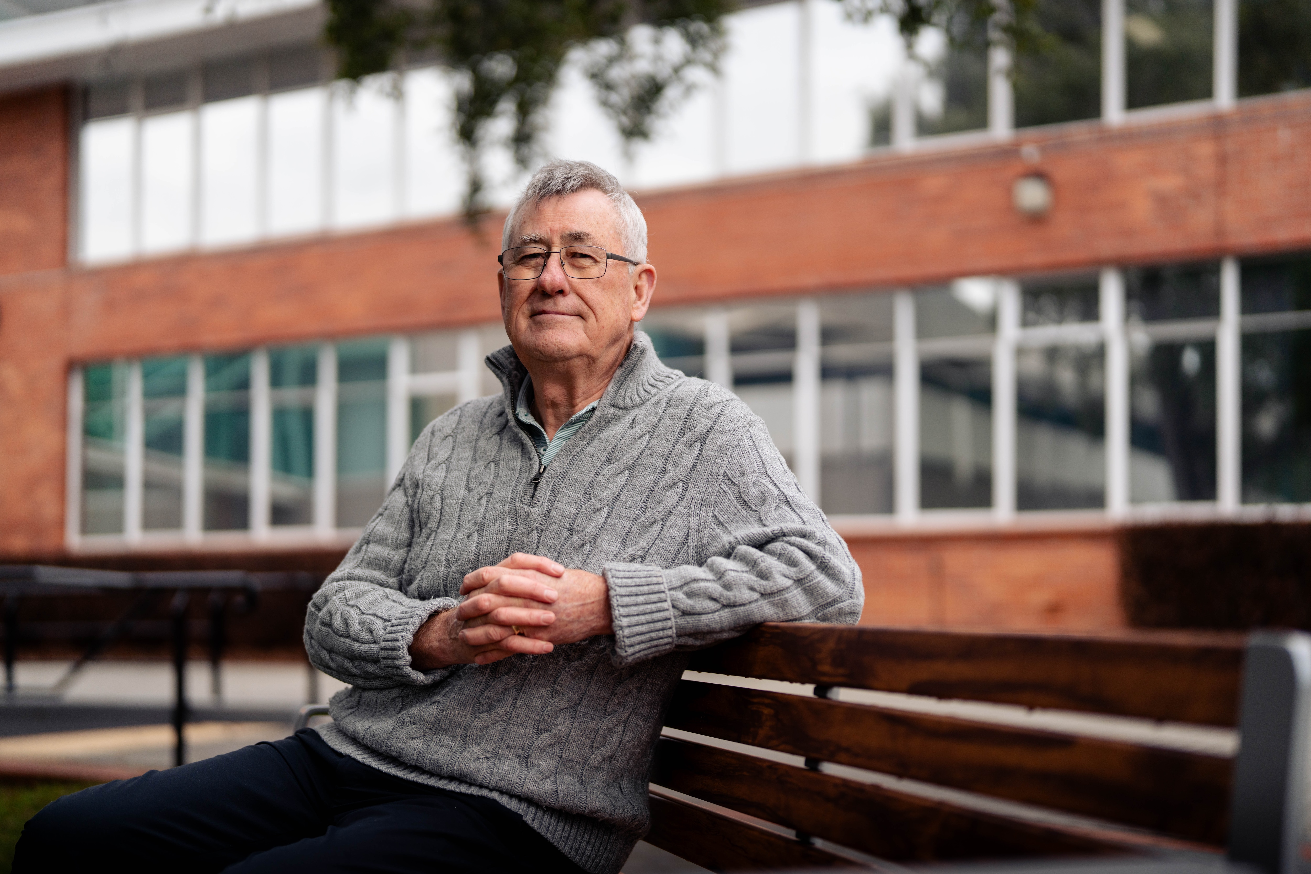 Brendan, wearing a grey quarter zip, sits at a park bench with his elbow resting on the top of the seat, smiling softly.