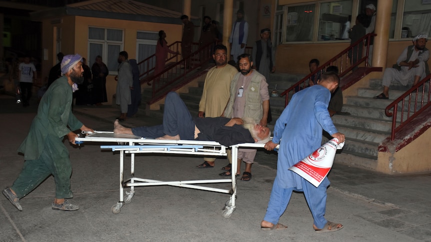 Two men in Middle Eastern garb wheel a bearded man on a stretcher outside a building at night